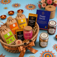 Handwoven Diwali gift basket tied with a copper ribbon holding dry fruit potlis, a jasmine candle and chai sachets, with two cookie jars, lit clay diyas and rangoli beside it