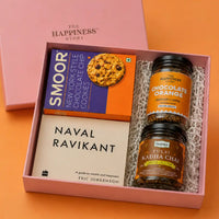 Top-down view of the open book gift hamper showing the two-column packing on white paper shred — cookie carton above the paperback on the left, coffee and chai jars stacked on the right