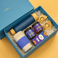 Open ocean blue Diwali gift box packed with a frosted glass bottle, a wheat straw sipper mug, two glass jars of loose leaf tea, a snack mix carton and two brass lotus diya holders on white paper shred