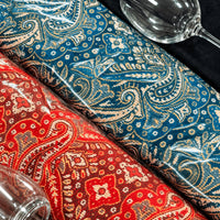 Close up of the woven paisley motif on the blue and red jamawar shawls, with the bowl and stem of a wine glass at the edge of the frame