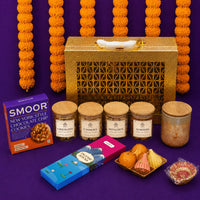 Gold Diwali gift case standing against marigold garlands with all its contents laid out in front — four jars, cookies, incense sticks, a scented candle, modak and ladoo candles and a hand-painted diya