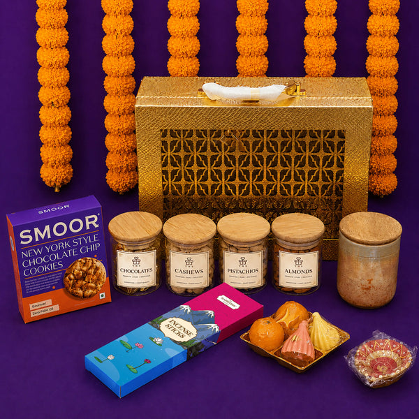 Gold Diwali gift case standing against marigold garlands with all its contents laid out in front — four jars, cookies, incense sticks, a scented candle, modak and ladoo candles and a hand-painted diya