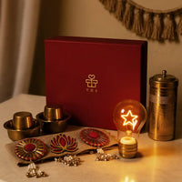 Styled Diwali still life with the star-filament bulb lit in its ribbed brass rechargeable base, brass dabara sets, coffee filter and jute toran in front of the upright gift box