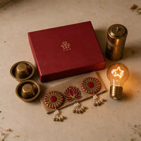 Flat-lay of the complete brass Diwali gift hamper on pale stone: closed red box, two dabara sets, brass coffee filter, jute toran and lit star bulb