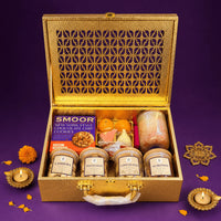 Open gold Diwali gift case filled with jars of cashews, pistachios, almonds and chocolates, a scented candle, modak and ladoo candles, cookies and incense, with lit brass diyas beside it