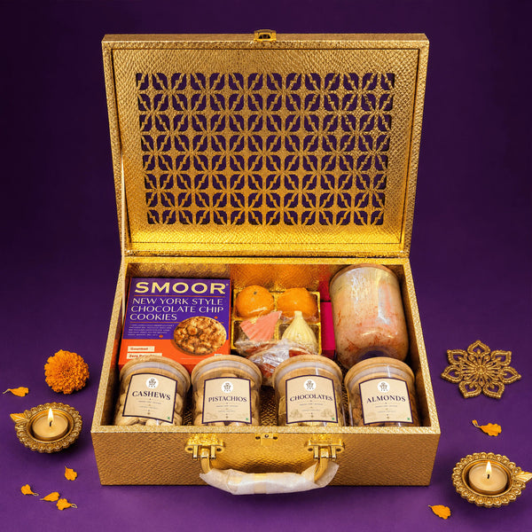 Open gold Diwali gift case filled with jars of cashews, pistachios, almonds and chocolates, a scented candle, modak and ladoo candles, cookies and incense, with lit brass diyas beside it