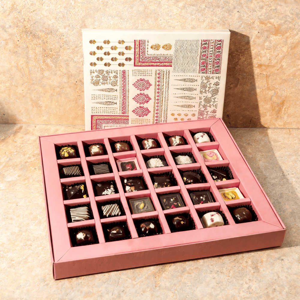 Open premium chocolate gift box with 25 assorted chocolates in a pink divided tray, block-printed lid standing behind