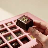 Hand lifting one 15 gram dark chocolate bonbon topped with pistachio and rose petal from the gift box