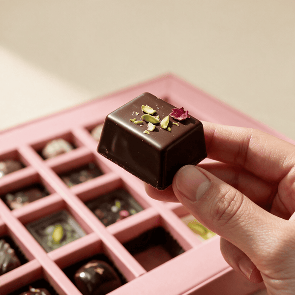 Hand lifting one 15 gram dark chocolate bonbon topped with pistachio and rose petal from the gift box