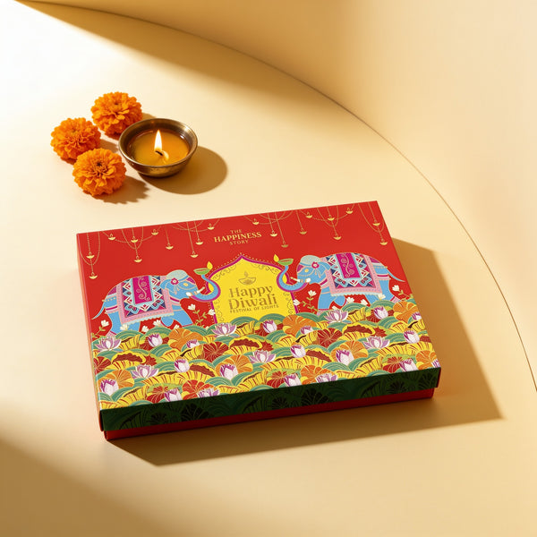 Closed red Subh Deepavali gift box with elephant and lotus artwork and Happy Diwali, Festival of Lights in gold, beside a brass diya and marigolds